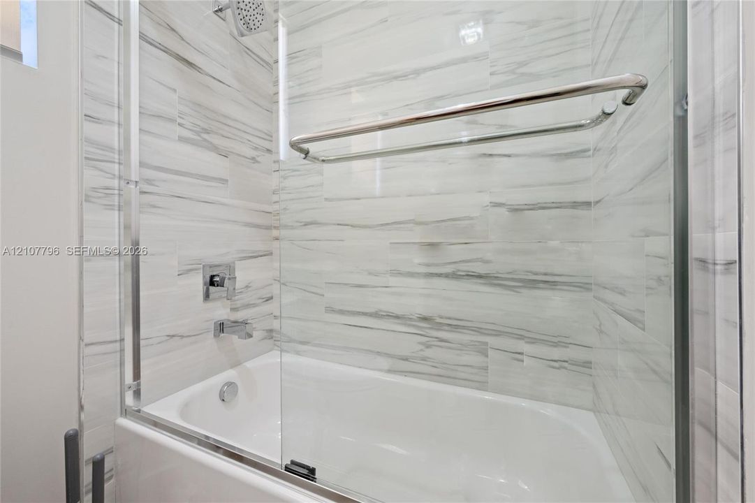 Bathtub featuring floor-to-ceiling stone-look tile surround, glass pivot door, wall-mounted chrome showerhead, and integrated chrome towel bar