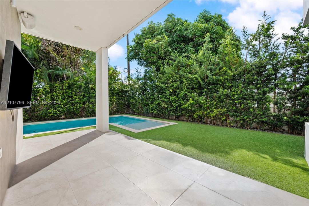 Covered patio with large-format tile flooring, in-ground swimming pool, synthetic turf lawn, and perimeter privacy hedging