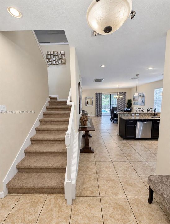 Carpeted staircase with white painted balustrade, tile flooring, kitchen featuring dark cabinetry and stainless steel appliances, and ceiling-mounted light fixtures