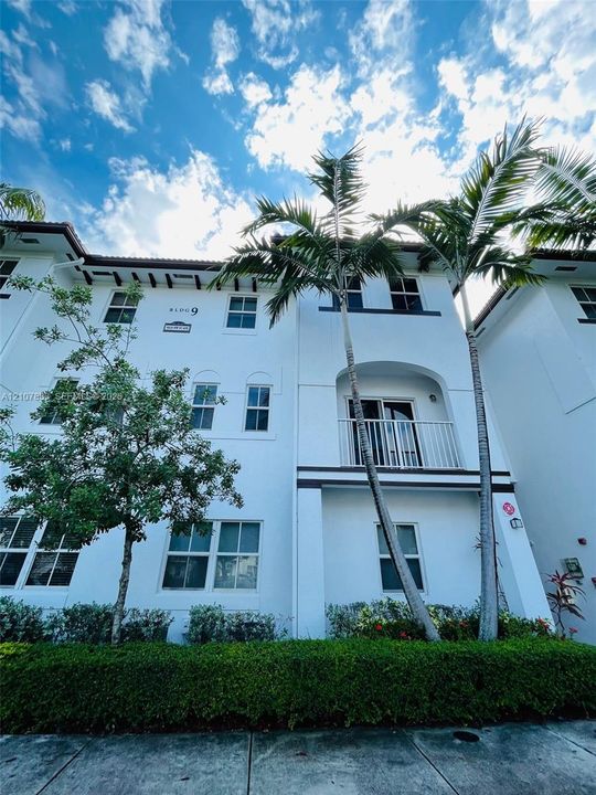 Multi-story building facade featuring white stucco finish, decorative bracket eaves, a private balcony with metal railing, and mature palm landscaping