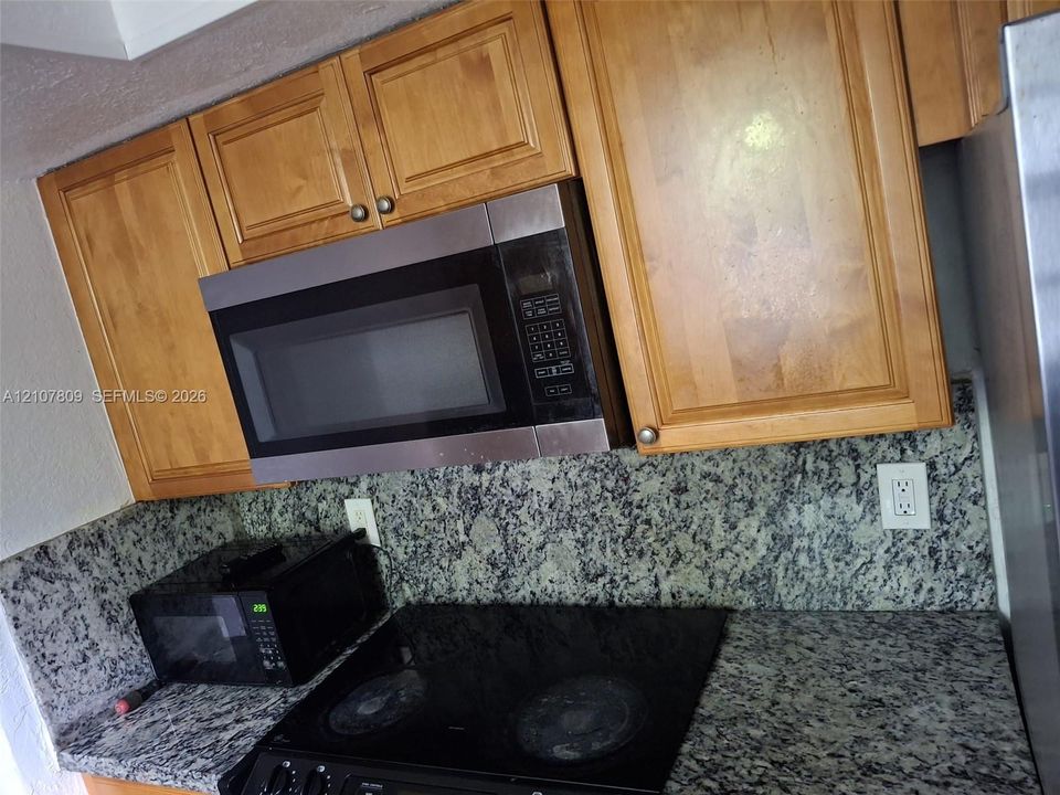 Natural wood cabinetry, speckled stone countertops, matching stone backsplash, stainless steel built-in microwave, and electric cooktop