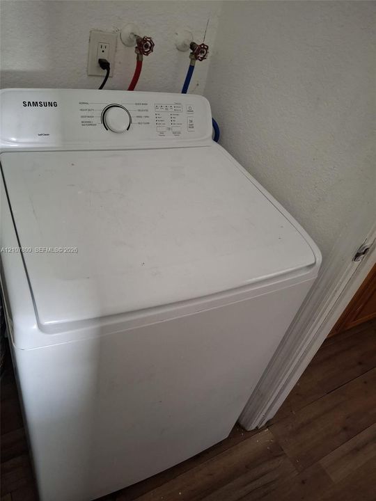 Top-loading Samsung washing machine with dedicated hot and cold water supply lines and wood-finish flooring