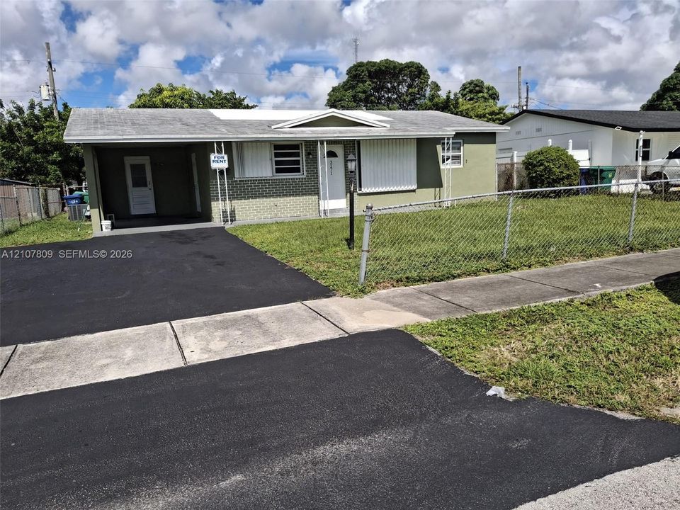 Single-story residence featuring a brick facade, covered carport, chain-link perimeter fencing, and an asphalt driveway