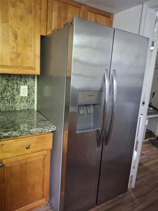 Stainless steel side-by-side refrigerator with exterior water and ice dispenser, natural wood-finish cabinetry, speckled stone-surface countertops, and wood-finish flooring