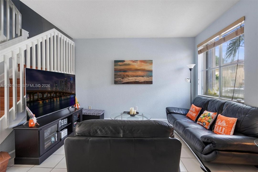Living area featuring ceramic tile flooring, a large window with blind, and a white staircase with wooden handrail