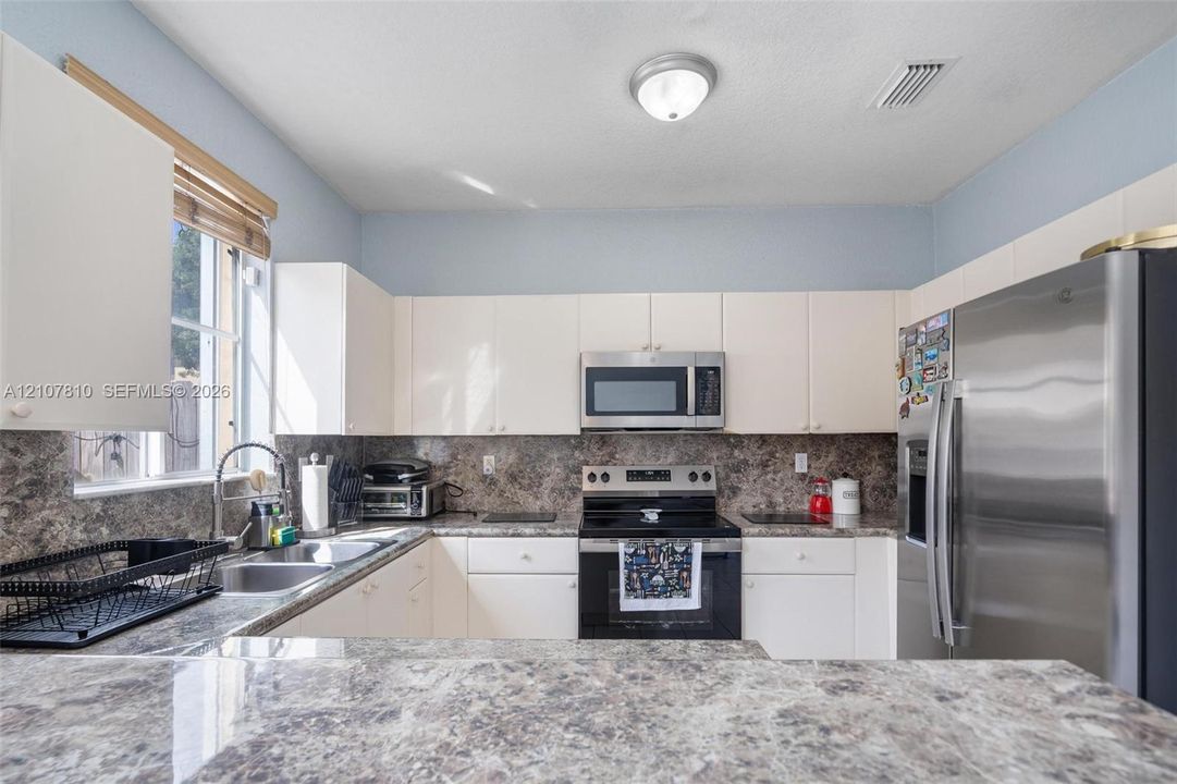 Kitchen featuring white cabinetry, stone-patterned countertops, stainless steel refrigerator, over-the-range microwave, and dual-basin sink with a gooseneck faucet