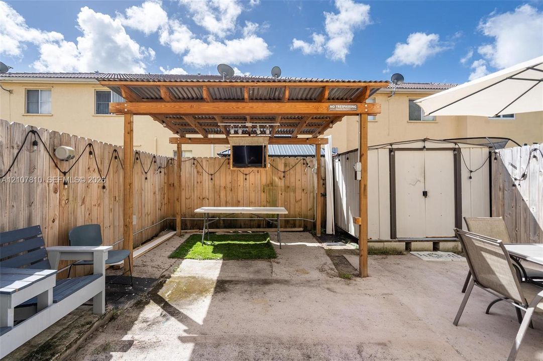 Private rear courtyard featuring a covered timber-framed pergola with a corrugated metal roof, concrete patio, perimeter wood fencing, and a detached storage structure. Outdoor furniture, BBQ Grill and Outside TV stays.