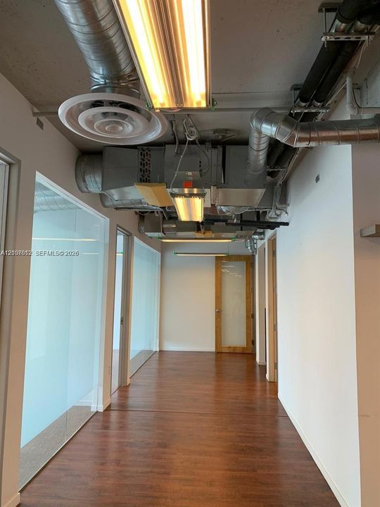 Interior corridor featuring wood-finish flooring, floor-to-ceiling glass wall partitions, exposed industrial-style ductwork, and linear overhead lighting
