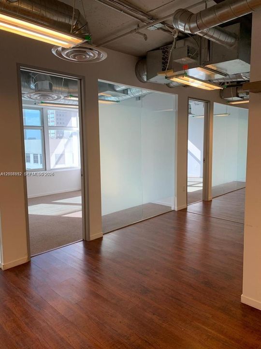 Exposed industrial ceiling with ductwork, wood-finish flooring, floor-to-ceiling glass partitions, and linear suspended lighting