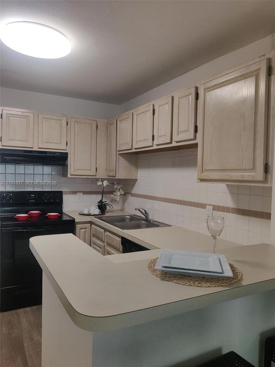Kitchen featuring light-toned cabinetry, dual-basin stainless steel sink, solid surface countertop with extended breakfast bar, tiled backsplash, and black appliances