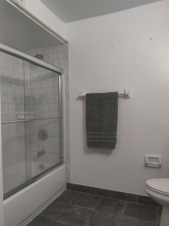 Shower-tub combination featuring a sliding glass enclosure, light-toned wall tile surround, dark slate-finish flooring, and chrome-finish plumbing fixtures
