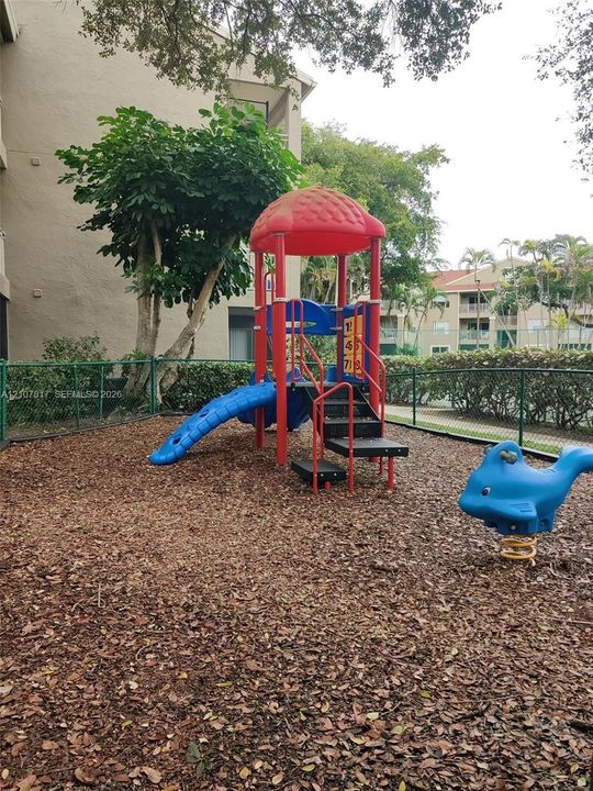 Outdoor recreational area featuring a multi-level climbing structure with slide, spring-mounted animal rider, wood chip surfacing, and perimeter chain-link fencing