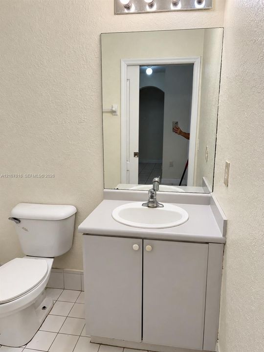 Vanity featuring solid-surface countertop with integrated basin, base cabinetry, full-length mirror, multi-bulb overhead light fixture, and ceramic tile flooring