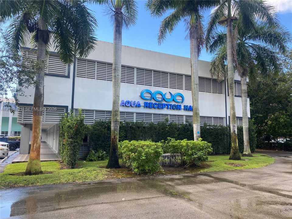 Two-story commercial building exterior featuring a concrete facade, horizontal window louvers, mature palm trees, and landscaped shrubbery