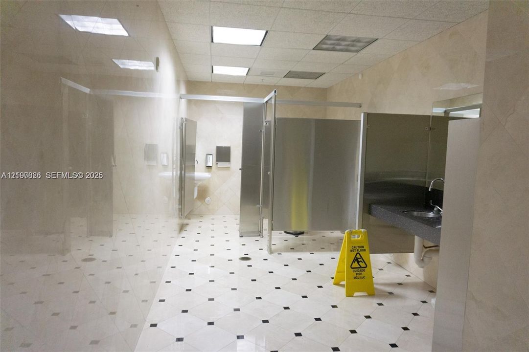 Restroom featuring floor-to-ceiling neutral wall tile, square-patterned tile flooring with dark inlays, stainless steel partitions, and a dark-toned vanity countertop with a drop-in sink