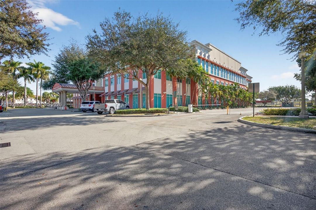 Commercial building exterior featuring a multi-story facade, covered porte-cochère, expansive paved surface parking, and mature palm and shade tree landscaping