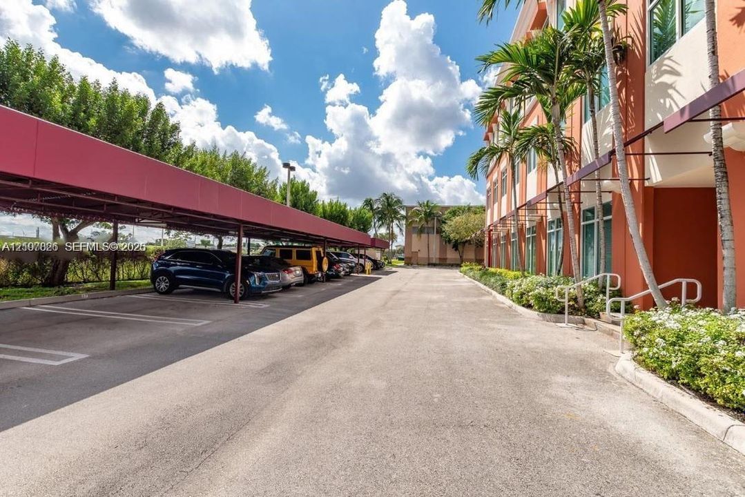 Asphalt parking area featuring covered vehicle shelters, multi-story building exterior with vertical windows, and manicured tropical landscaping with palm trees