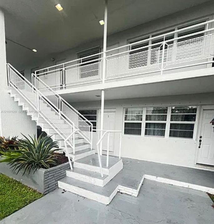Two-story exterior featuring white-painted masonry, metal staircases with diamond-patterned railings, a paved ground-level entry, and a concrete planter box