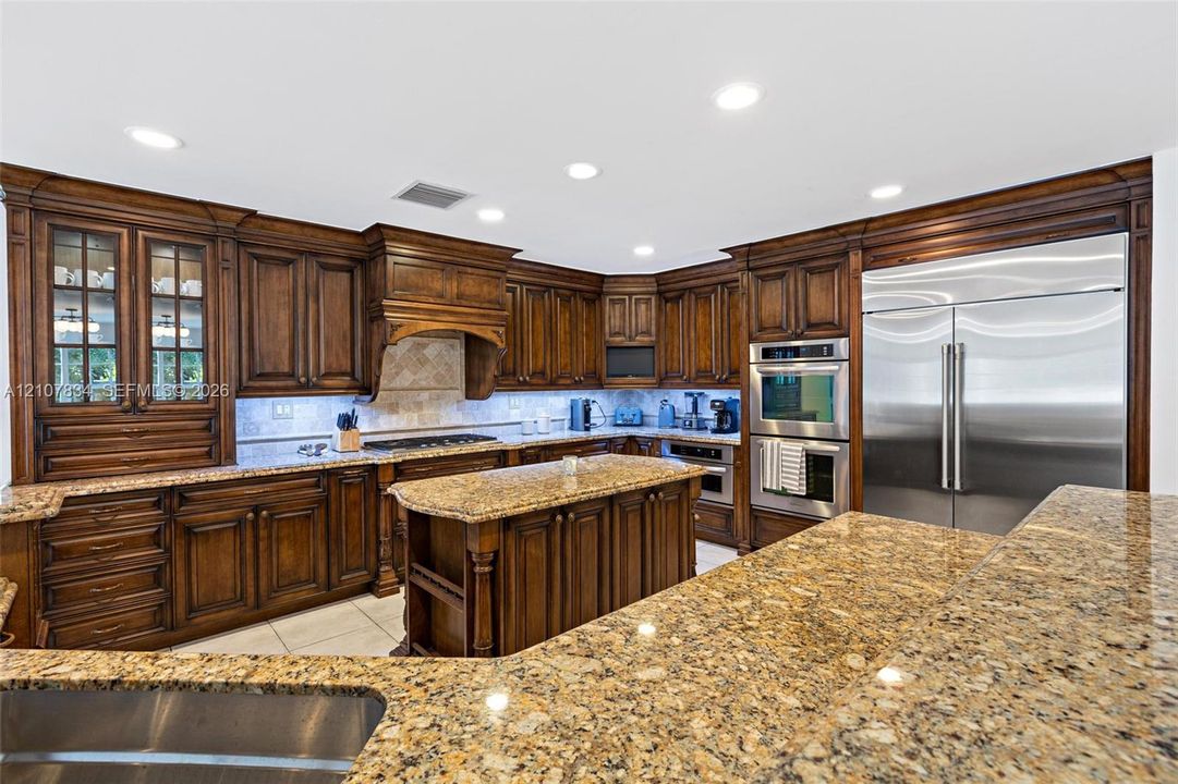 Kitchen featuring custom stained-wood cabinetry with glass-front display panels, natural stone countertops, a central island, recessed lighting, and integrated stainless steel double wall ovens and refrigerator