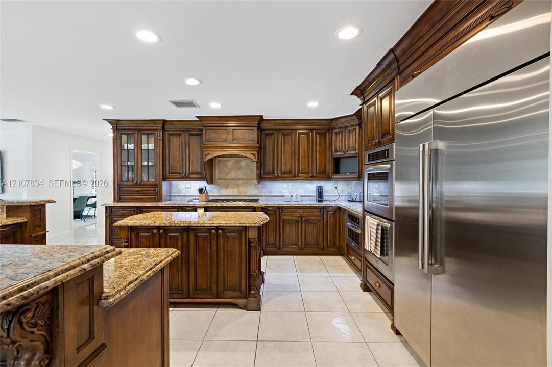 Kitchen featuring stained wood cabinetry, stone countertops, integrated stainless steel appliances, a central island with decorative corbels, and ceramic tile flooring