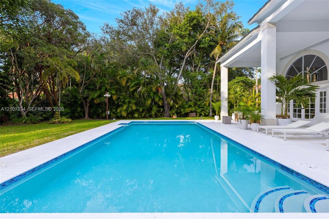 Rectangular swimming pool with tiled coping and tiered entry steps, complemented by an expansive stone-paved pool deck and a covered patio supported by structural columns