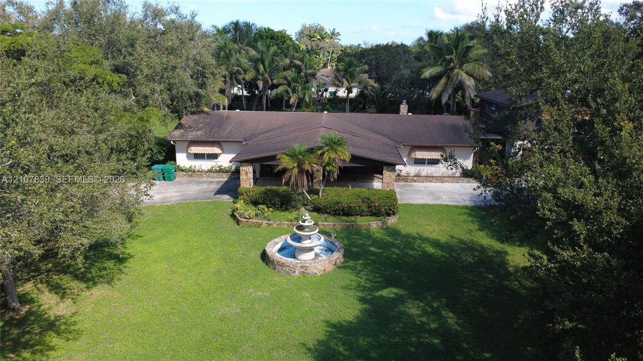 Single-story residence featuring a covered portico, stone-accented facade, central tiered water fountain, and expansive lawn surrounded by mature trees
