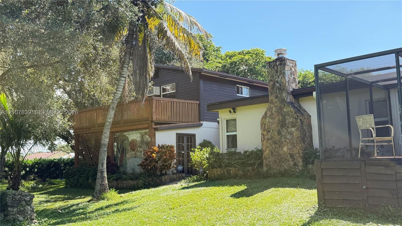 Multi-level exterior featuring a wood-railing deck, exterior stone chimney, screened-in enclosure, and mature palm landscaping