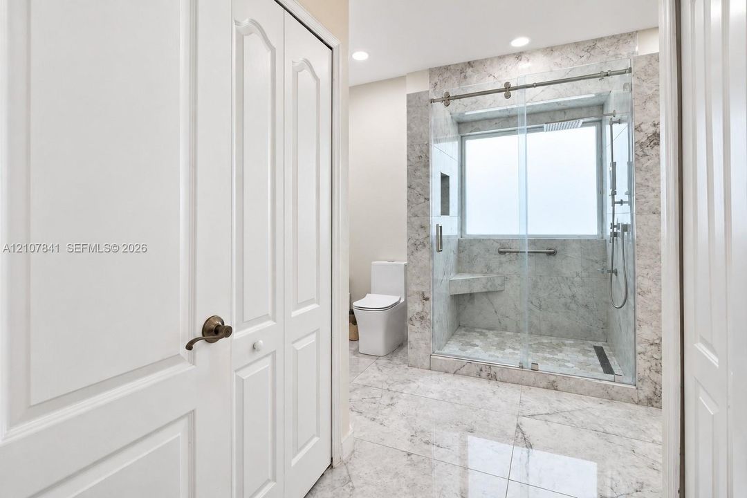 Walk-in shower with stone-finish tile surround, integrated bench seat, and sliding glass enclosure, complemented by large-format floor tiling and white dual-panel doors