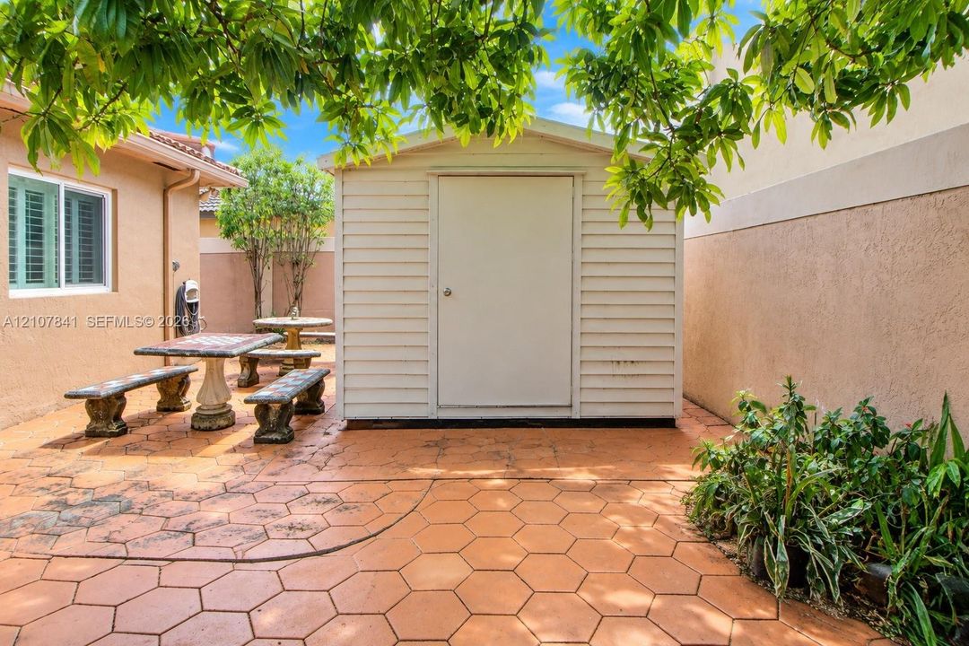 Exterior space featuring a neutral-toned storage shed, hexagonal paver hardscaping, and a stucco-finished wall