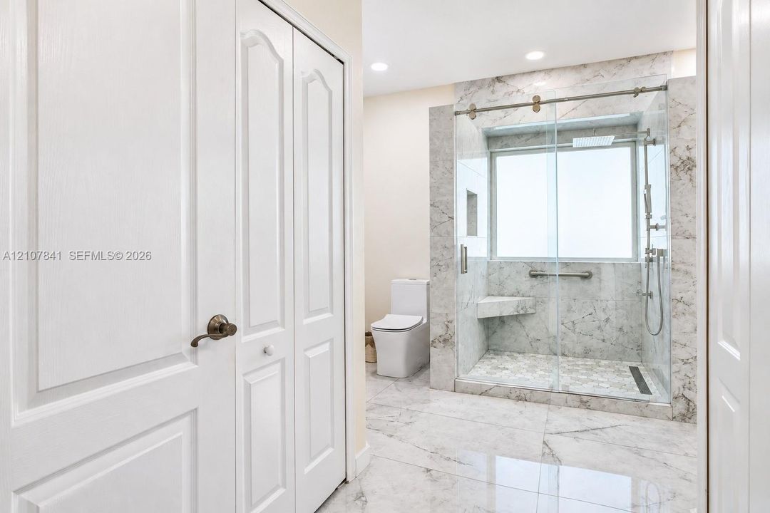 Walk-in shower with stone-finish tiling, integrated bench seat, glass sliding door, and wall-mounted shower system, complemented by stone-finish flooring and recessed lighting