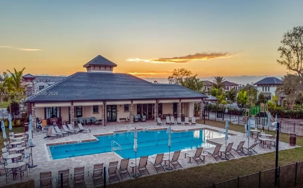 Community recreation center featuring a rectangular swimming pool with paved decking, a covered veranda, stone pillar accents, and a multi-sloped roofline