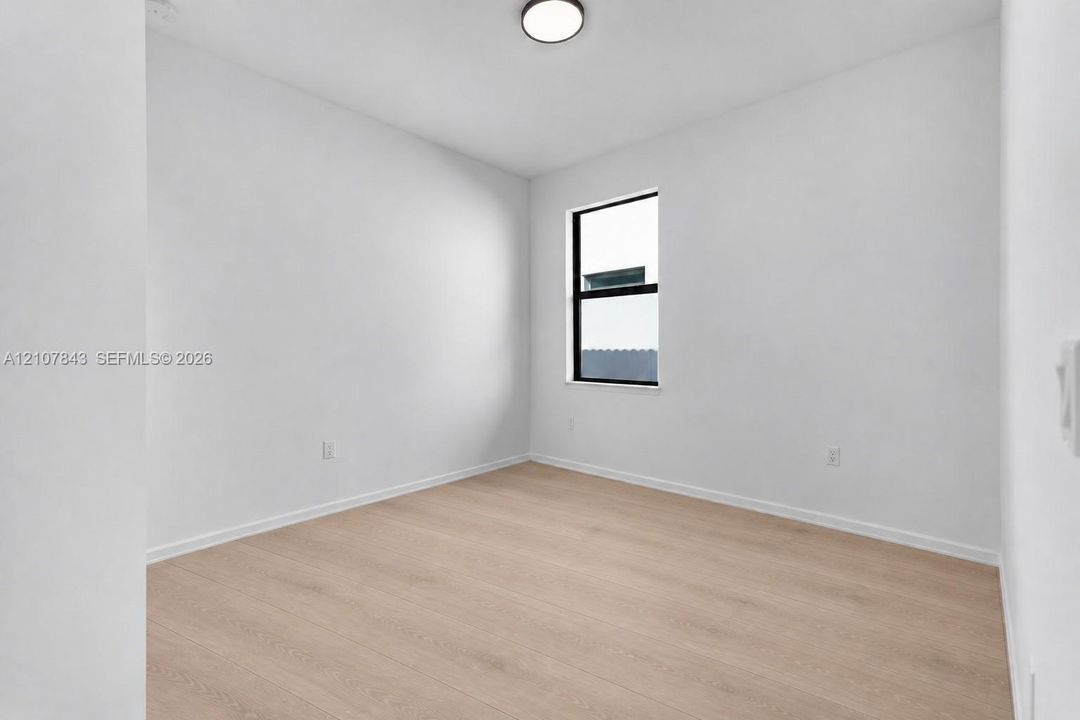 Neutral-toned interior space featuring wood-finish flooring, crisp white wall finishes, and a single-pane window with a black frame, completed by a minimalist flush-mount ceiling light fixture