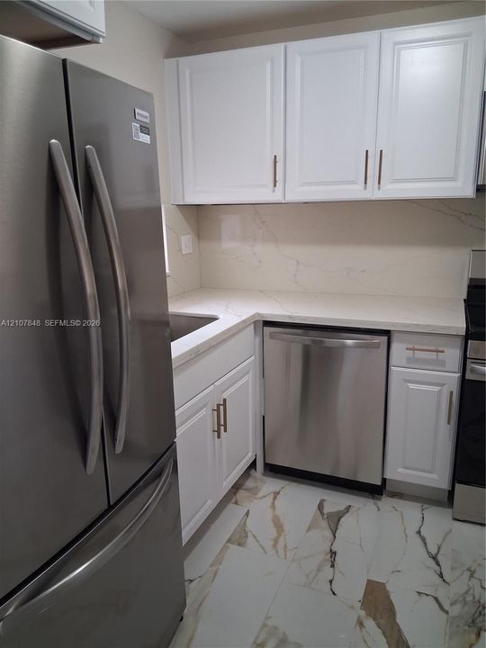 Kitchen featuring white shaker-style cabinetry, stainless steel appliances, solid surface countertops, a full-height stone-finish backsplash, and large-format porcelain tile flooring