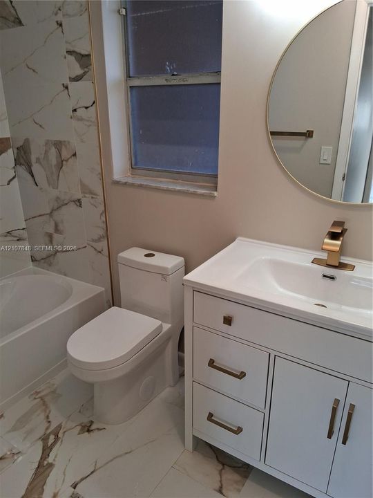 Bathroom featuring a white vanity with metallic hardware, integrated sink, gold-toned faucet, round wall-mounted mirror, and stone-patterned tile flooring and shower surround