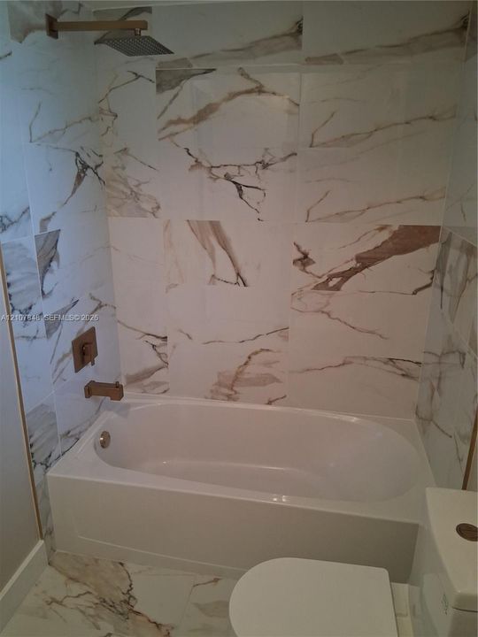Full-height, large-format stone-patterned wall tile, brass-finish rainfall showerhead, wall-mounted brass-finish faucet, and integrated soaking tub