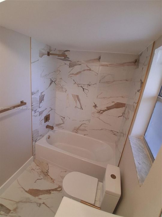Full-height tile shower surround with integrated bathtub, patterned stone-finish floor tiling, wall-mounted brushed gold-tone shower hardware, and custom stone-surround window sill