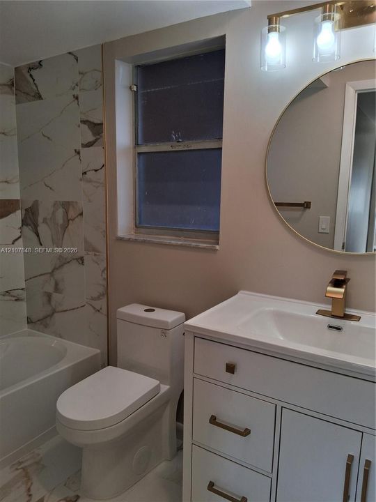 White vanity with a rectangular sink, brushed gold faucet, and brass-tone hardware, paired with a round mirror, gold-toned light fixture, and stone-finish wall tile surrounding the bathtub