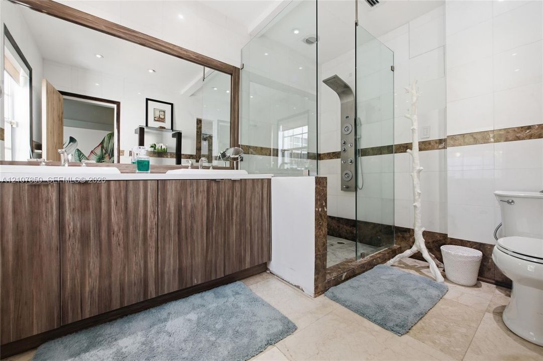 Dual-sink vanity with wood-finish cabinetry, full-width mirror, walk-in shower with frameless glass enclosure, and stone-tile flooring