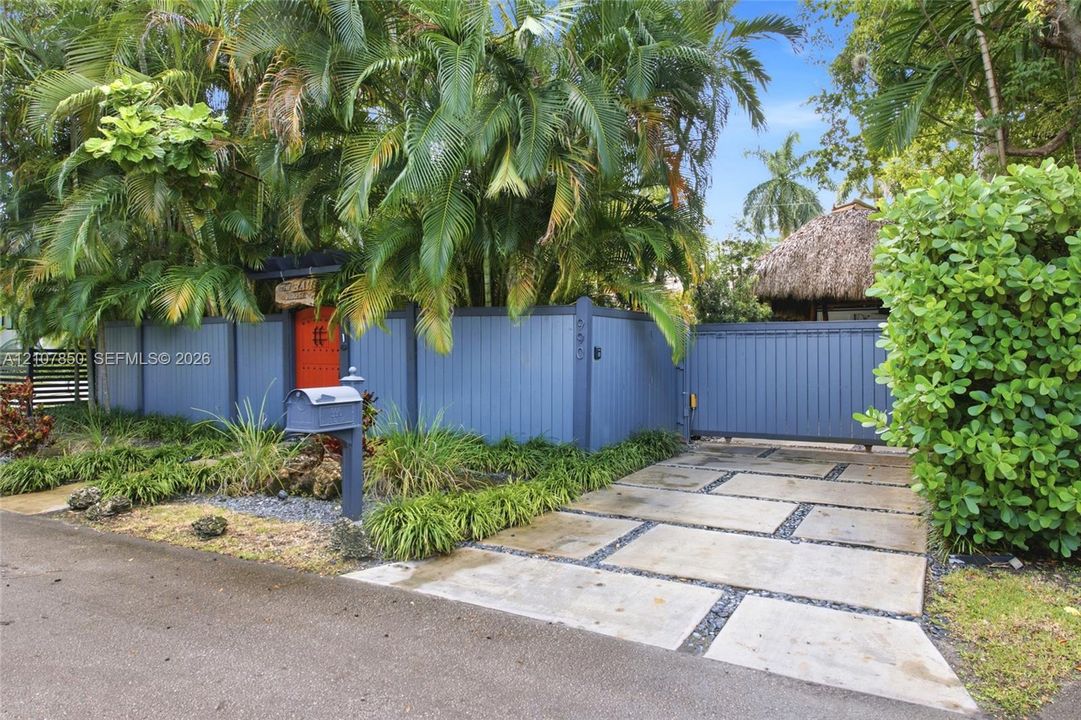 Private gated entry featuring a custom blue vertical-board fence, concrete paver driveway with crushed stone detailing, and mature tropical landscaping