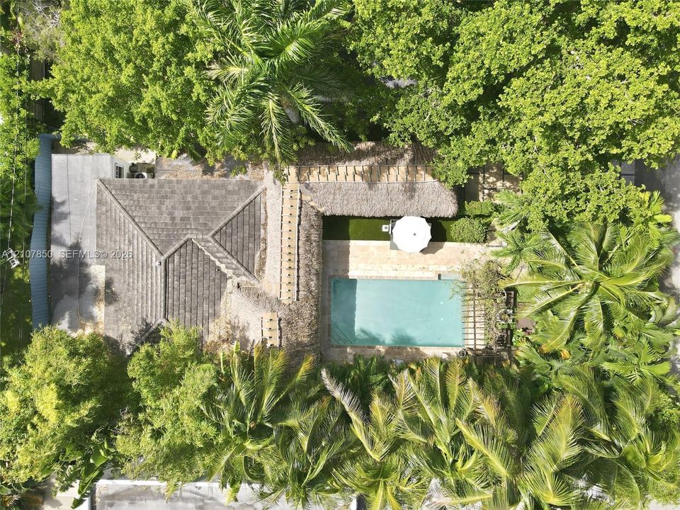 Aerial perspective featuring a rectangular swimming pool, stone-paved patio, thatched roof structure, and lush mature trees