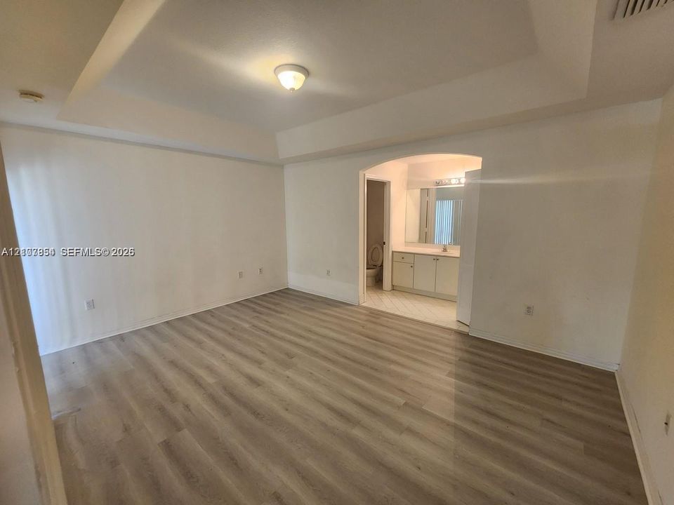 Tray ceiling, wood-finish flooring, and arched entryway leading to a bathroom with a neutral-toned vanity and full-width vanity mirror