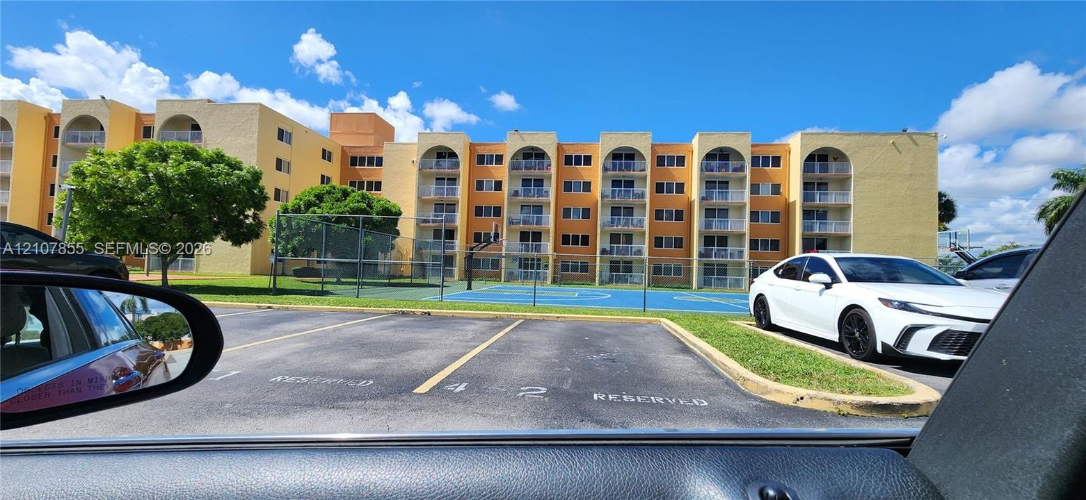Multi-story building exterior featuring arched private balconies, a hard-surfaced sports court, and designated parking areas