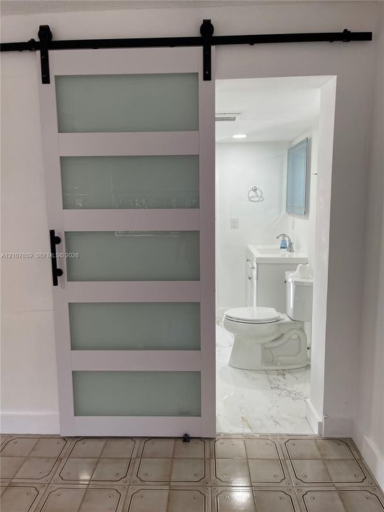 Sliding barn door featuring frosted glass panels and black hardware, opening into a bathroom with a white vanity, stone-finish floor tile, and chrome fixtures