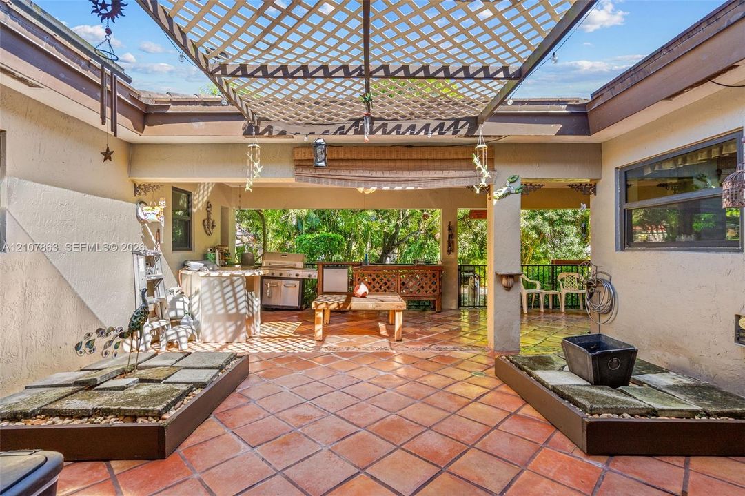 Covered outdoor patio featuring tile flooring, built-in outdoor kitchen, lattice overhead structure, and dual raised stone planters