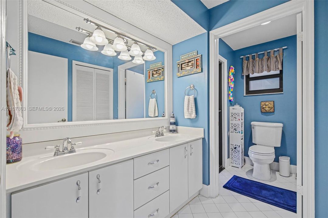Dual-sink vanity featuring a solid surface countertop, white cabinetry with polished hardware, a large wall-mounted mirror, and a multi-bulb light fixture