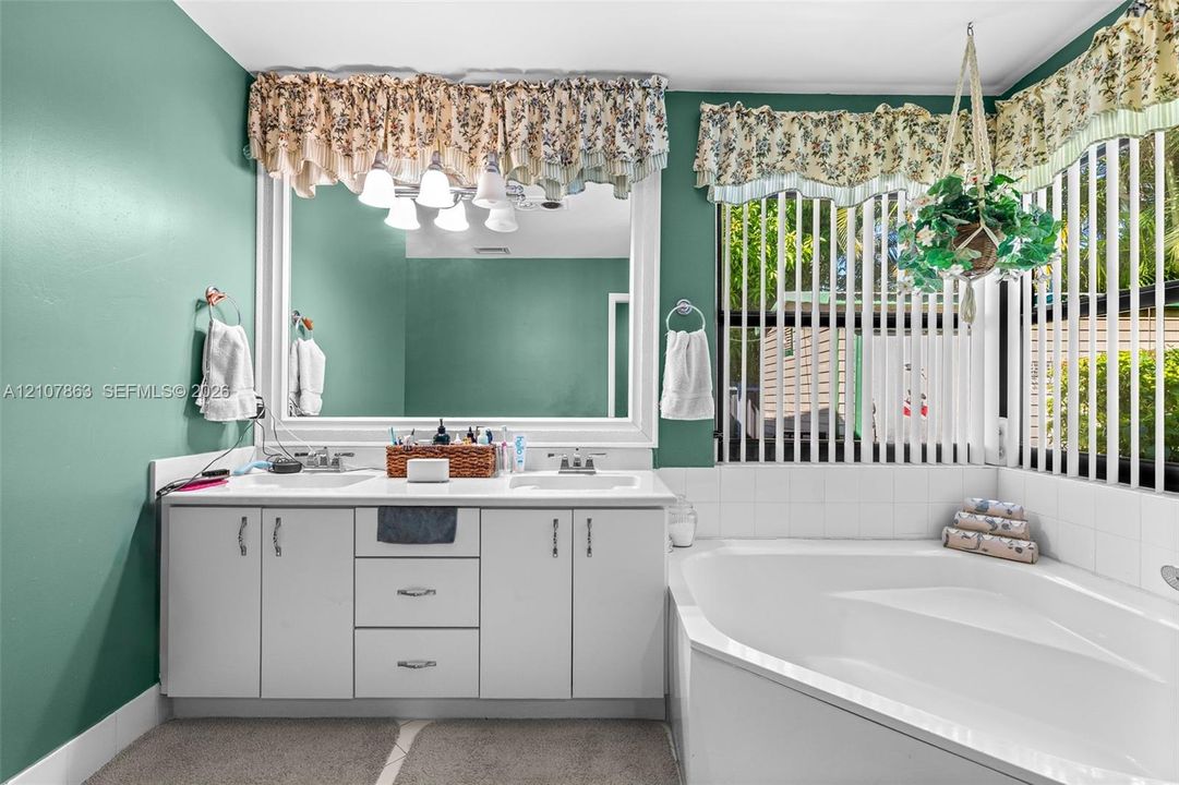 Double vanity featuring white cabinetry, a solid-surface countertop with integrated dual sinks, a large wall-mounted mirror, and a corner soaking tub with tiled surround