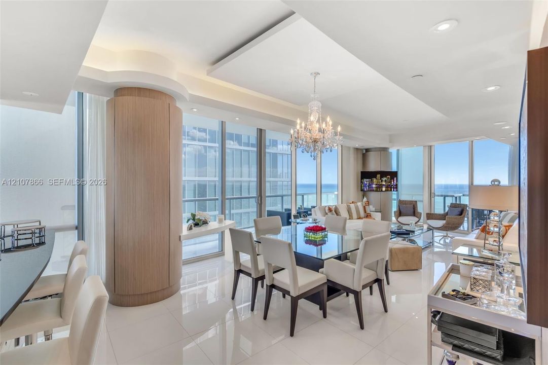 Open-concept interior featuring polished tile flooring, tray ceilings with recessed lighting, floor-to-ceiling glass windows, a circular wood-finish architectural column, and a crystal chandelier