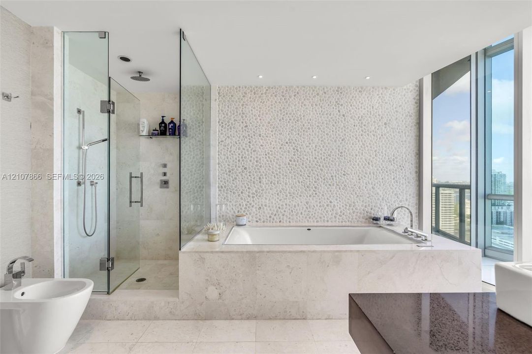 Bathroom featuring a built-in soaking tub, glass-enclosed walk-in shower with a rainfall showerhead, stone-tile flooring, textured stone-accent wall, and floor-to-ceiling windows
