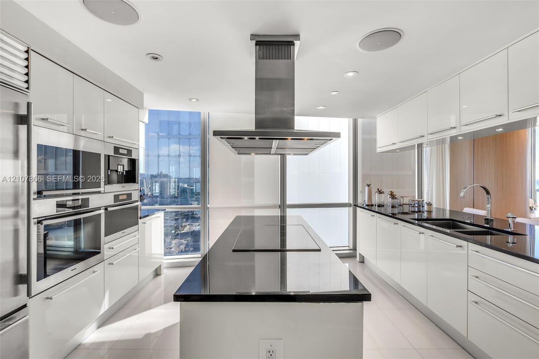 Modern kitchen featuring high-gloss white cabinetry, a center island with a dark solid-surface countertop, stainless steel integrated wall ovens, a ceiling-mounted stainless steel vent hood, and floor-to-ceiling windows
