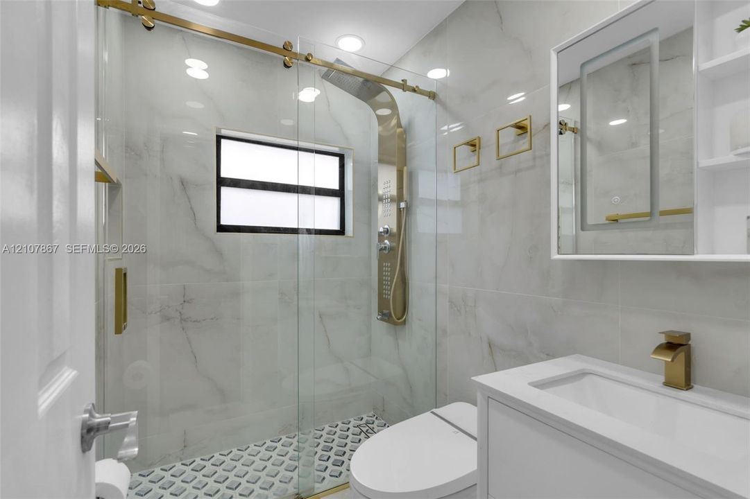 Walk-in shower with floor-to-ceiling tile, brass-finish rainfall shower panel, sliding glass door, geometric floor tile, and vanity with rectangular basin and brass-finish faucet